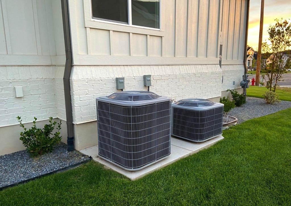 Two outdoor HVAC air conditioning units installed beside a residential home on concrete pads, surrounded by lawn and landscaping.